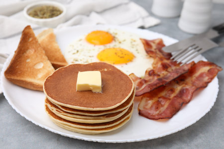 Tasty pancakes served with fried eggs and bacon on gray table, closeup