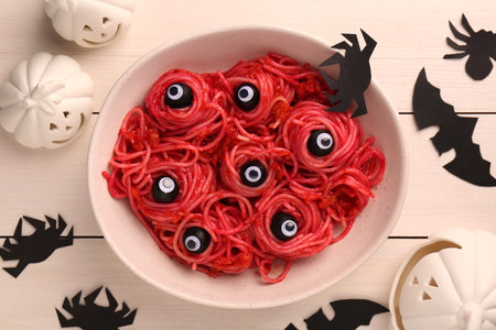 Red pasta with decorative eyes and olives in bowl served on white wooden table, flat lay. Halloween foodの写真素材