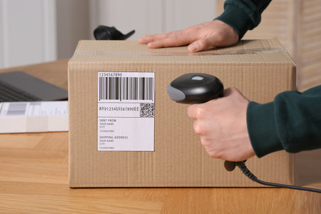 Seller with scanner reading parcel barcode at table in office, closeup. Online store