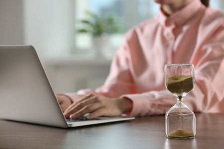 Hourglass with flowing sand on wooden table, selective focus. Man using laptop indoors