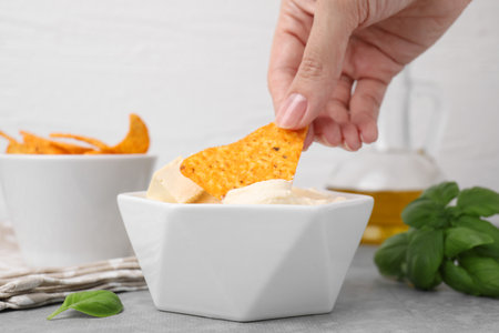 Woman dipping nachos chip into delicious tofu sauce at gray table, closeup