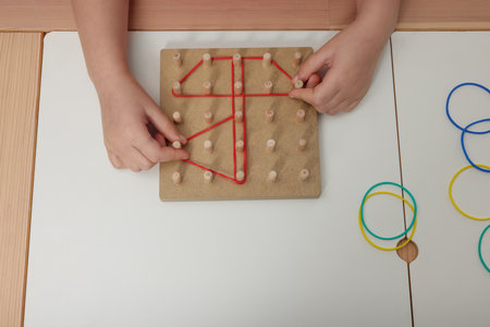 Motor skills development. Girl playing with geoboard and rubber bands at white table, closeupの写真素材
