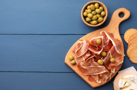 Slices of tasty cured ham, olives and cheese on blue wooden table, flat lay. Space for textの写真素材