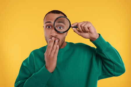Surprised man looking through magnifier glass on orange backgroundの写真素材