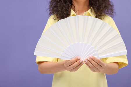 Woman holding hand fan on purple background, closeup. Space for textの写真素材