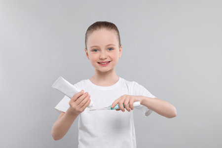 Happy girl squeezing toothpaste from tube onto electric toothbrush on light gray background