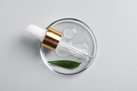 Petri dish with sample of cosmetic oil, pipette and green leaf on white background, top viewの写真素材