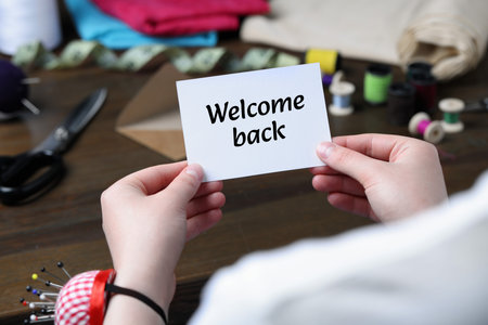 Seamstress holding paper note with phrase Welcome Back at table in atelier, closeupの写真素材