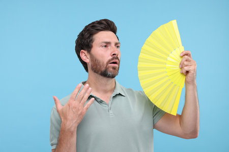 Unhappy man with hand fan suffering from heat on light blue backgroundの写真素材