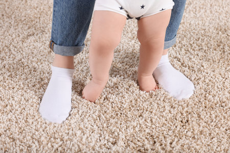 Mother supporting her baby son while he learns to walk on carpet at home, closeup