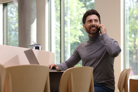 Handsome man talking on smartphone while using laptop at table in cafeの写真素材