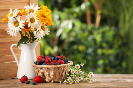 Wicker bowl with different fresh ripe berries and beautiful flowers on wooden table outdoors, space for textの写真素材