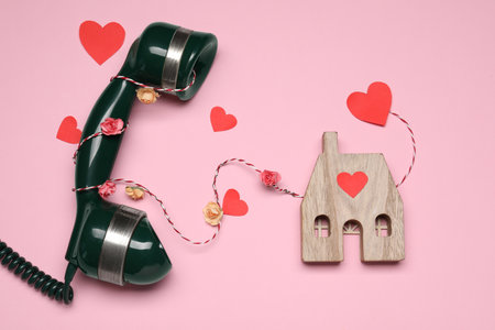Decorative cord and paper hearts between telephone receiver and wooden house model on pink background symbolizing connection in long-distance relationship, flat layの写真素材