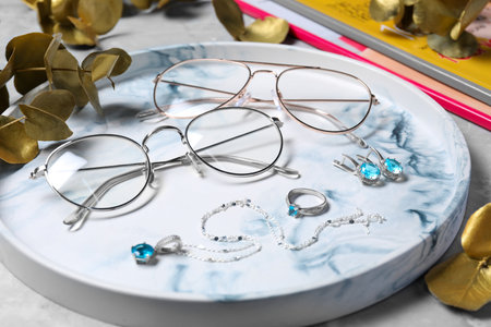 Glasses in stylish frames, leaves and jewelry on gray table