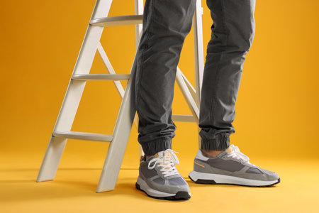 Man wearing stylish sneakers near white ladder on yellow background, closeup