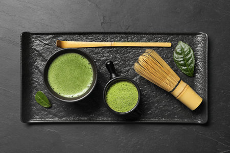 Cup of fresh matcha tea, bamboo whiskey, spoon and green powder on black textured table, top view