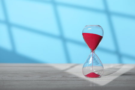 Hourglass with red flowing sand on white wooden table against light blue background, space for text