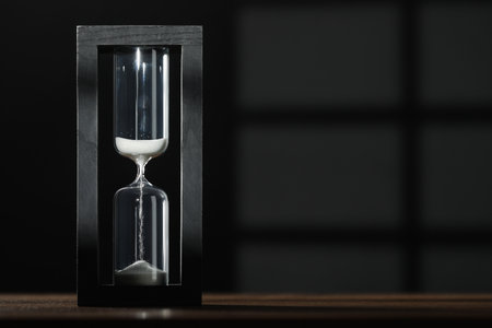 Hourglass with flowing sand on wooden table against black background, space for textの写真素材