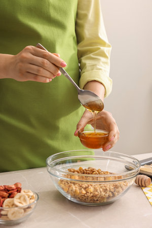 Making granola. Woman adding honey into bowl with mixture of oat flakes and other ingredients at light marble table in kitchen, closeupの写真素材