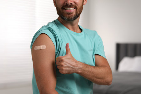 Man with sticking plaster on arm after vaccination showing thumbs up in bedroom, closeupの写真素材