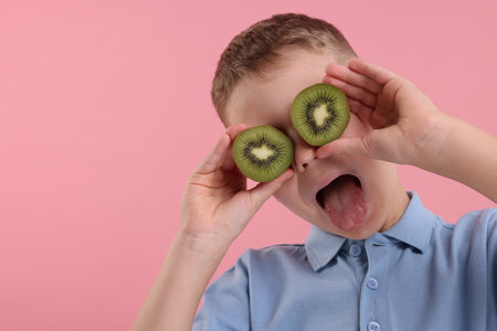 Emotional boy covering eyes with halves of fresh kiwi and showing tongue on pink background, space for textの写真素材