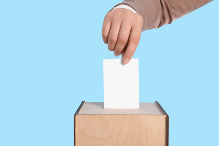 Woman putting her vote into ballot box on light blue background, closeup. Space for text
