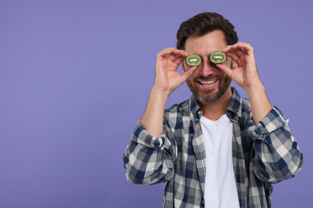Man holding halves of kiwi near his eyes on purple background, space for textの写真素材