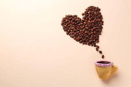 Coffee beans spilled from edible biscuit cup in shape of heart on beige background, top view. Space for textの写真素材