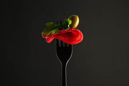 Fork with tasty slice of salami, olive and parsley on black background