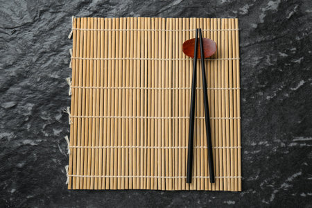Bamboo mat with pair of chopsticks and rest on black textured table, top viewの写真素材