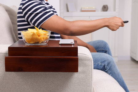 Nacho chips and smartphone on sofa armrest wooden table. Man holding remote control at home, closeupの写真素材