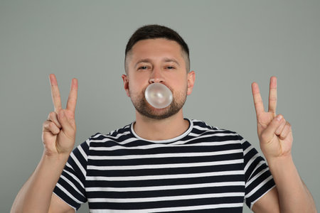 Handsome man blowing bubble gum and showing peace gesture on light grey backgroundの写真素材