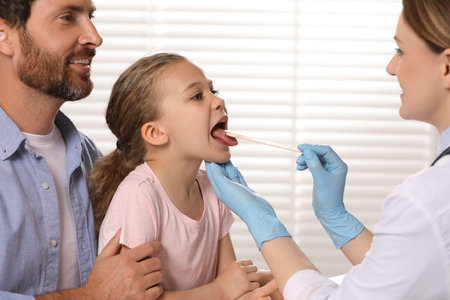 Doctor examining girl`s oral cavity with tongue depressor near her father indoorsの写真素材