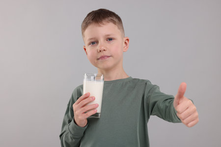 Cute boy with glass of fresh milk showing thumb up on light grey backgroundの写真素材