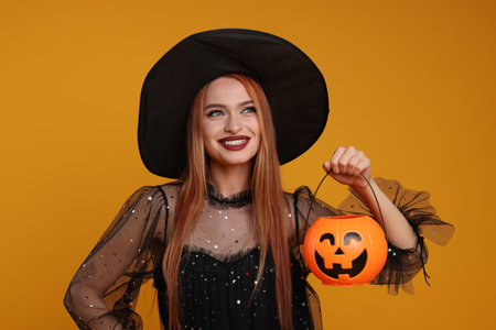 Happy young woman in scary witch costume with pumpkin bucket on orange background. Halloween celebrationの写真素材