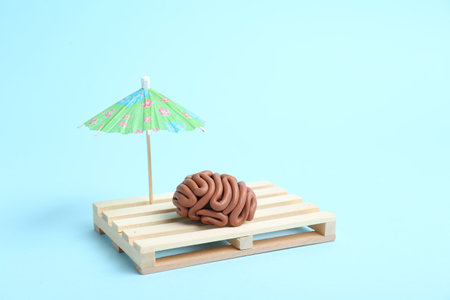 Brain made of plasticine on mini wooden sunbed under umbrella against light blue background, space...