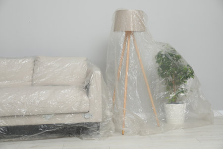 Sofa, lamp and houseplant covered with plastic film near light gray wall indoorsの写真素材