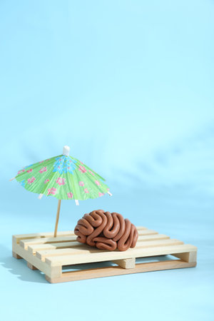 Brain made of plasticine on mini wooden sunbed under umbrella against light blue background, space for textの写真素材