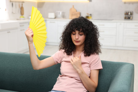 Young woman waving yellow hand fan to cool herself on sofa at homeの写真素材