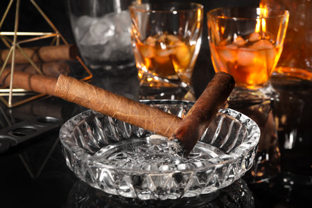 Smoldering cigars, ashtray and whiskey on black mirror surface, closeup