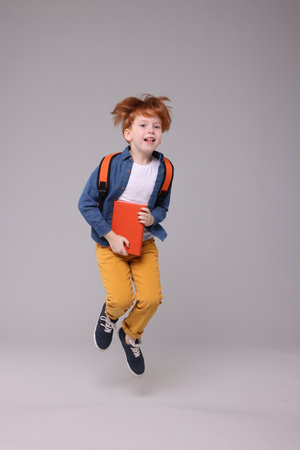 Happy schoolboy with backpack and book jumping on grey backgroundの写真素材