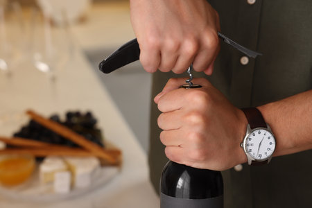 Man opening wine bottle with corkscrew indoors, closeup. Space for text