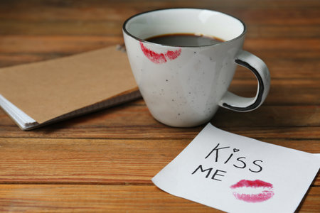 Notebook, sticky note with phrase Kiss Me, lipstick mark and cup of coffee on wooden tableの写真素材