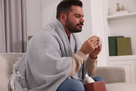 Sick man wrapped in blanket with tissues on sofa at home. Cold symptomsの写真素材