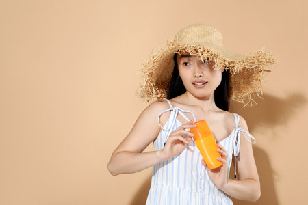 Beautiful young woman in straw hat holding sun protection cream on beige background, space for text
