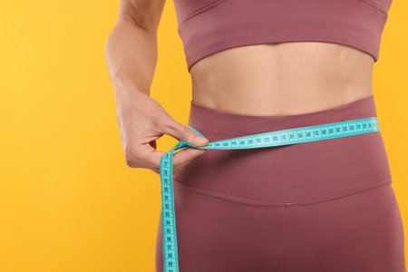 Slim woman measuring waist with tape on yellow background, closeup. Weight loss