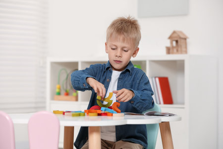 Cute little boy playing with colorful wooden pieces at white table indoors. Child's toy