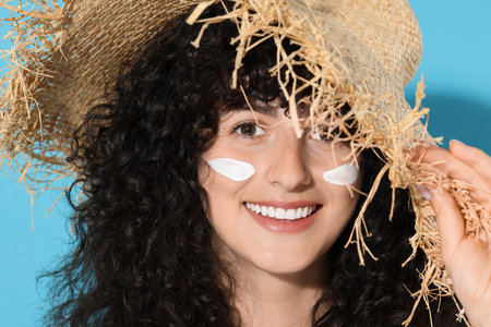 Beautiful young woman in straw hat with sun protection cream on her face against light blue...