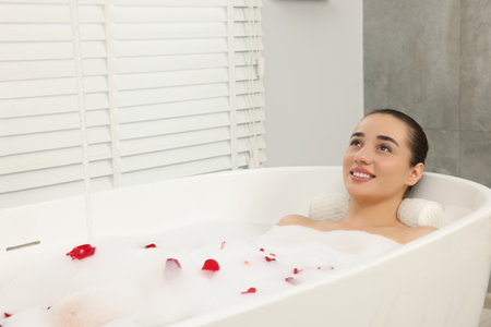Happy woman taking bath in tub with foam and rose petals indoorsの写真素材