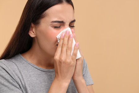 Suffering from allergy. Young woman blowing her nose in tissue on beige background, closeup. Space for textの写真素材